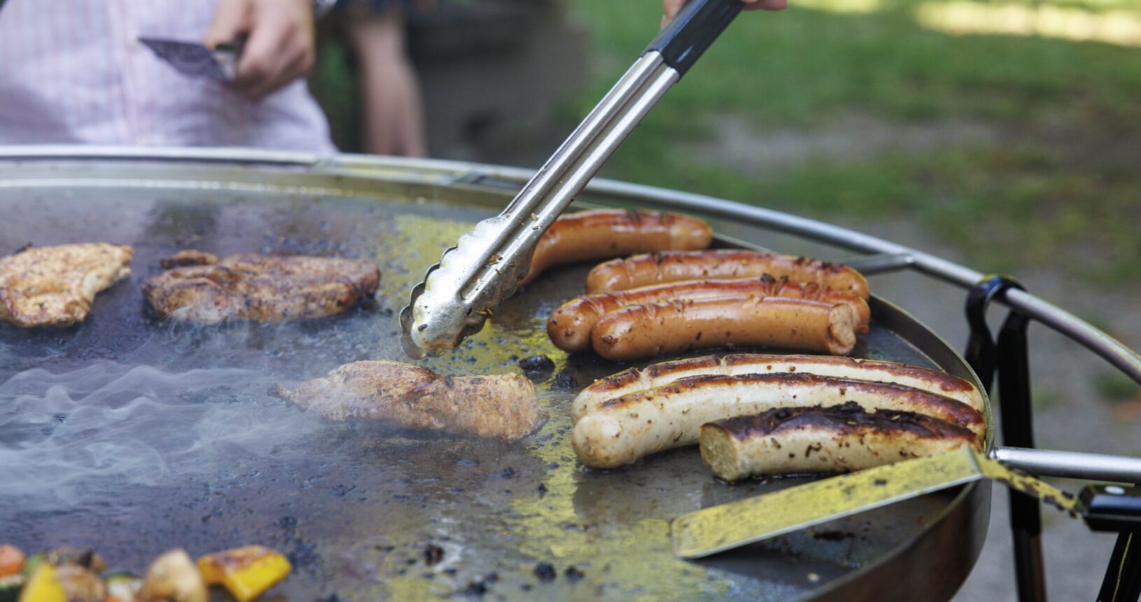 Eine Nahaufnahme eines runden Grills im Freien, auf dem Würstchen, Hähnchen und Gemüse brutzeln. Bei dieser Teambuilding-Veranstaltung benutzt eine Person eine Zange, um das Essen zu wenden, während eine andere in der Nähe steht und Rauch vom Grill aufsteigt.