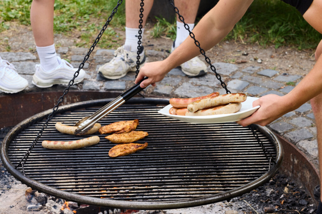 Ein Grillrost in Nahaufnahme. Man sieht wie fertige Würstchen mit einer Grillzange auf einen großen Teller genommen werden.