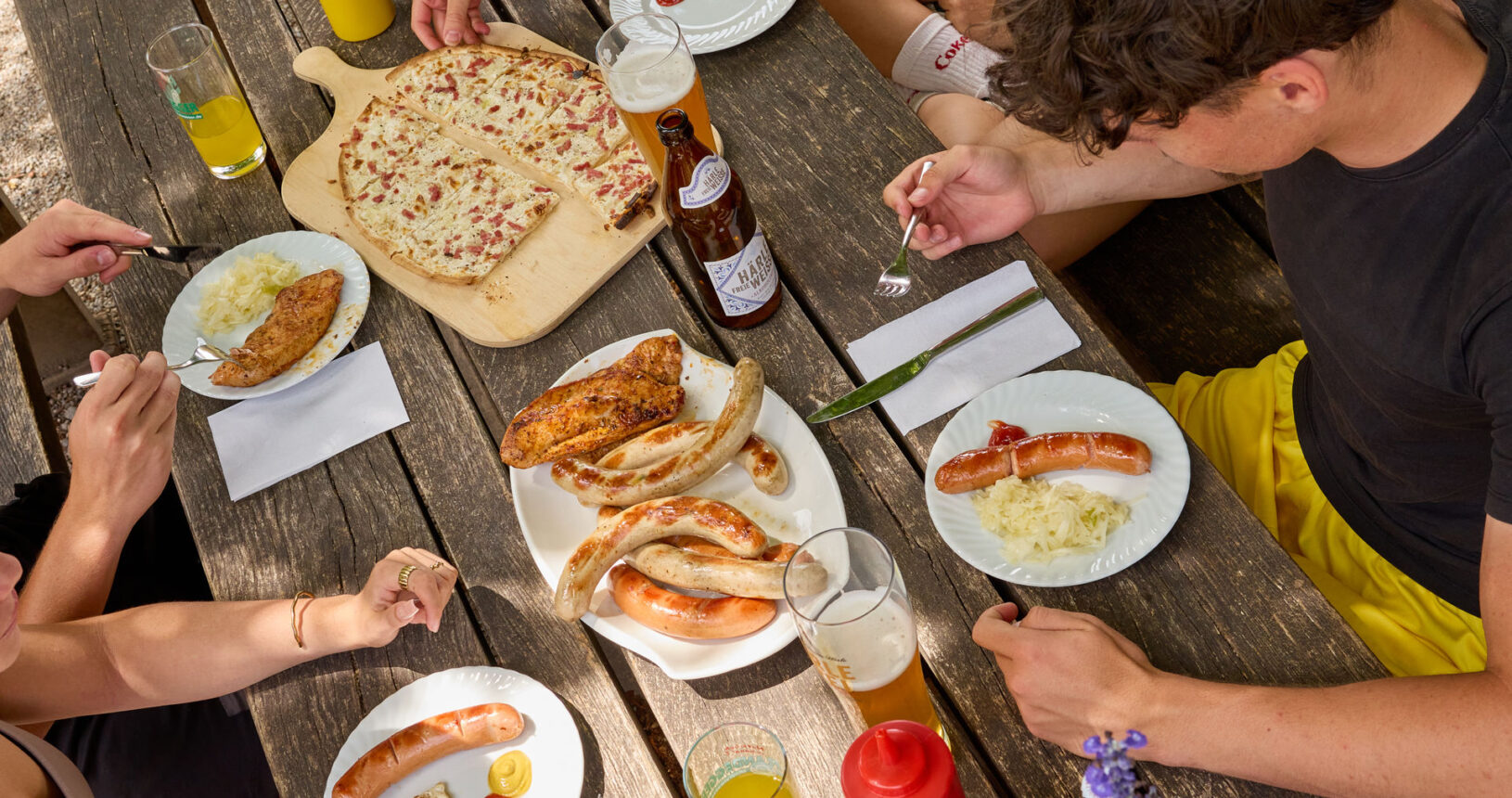 An einem Tisch sitzen mehrere Leute und essen. Man sieht unterschiedliche Speisen vom Grill sowie einen Flammkuchen und kühle Getränke.