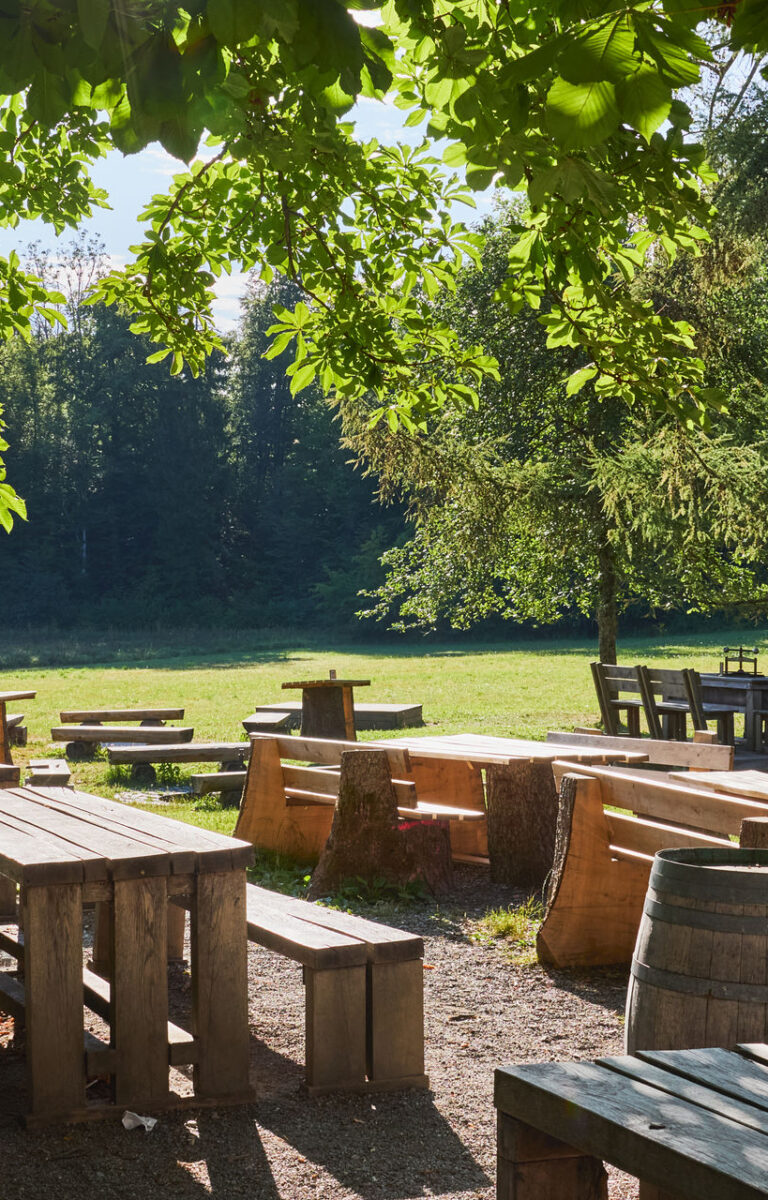 Ein rustikaler Biergarten mit massiven Holztischen, der in den frühen Morgenstunden von der Sonne angestrahlt wird. Im Hintergrund erkennt man eine große Wiese.