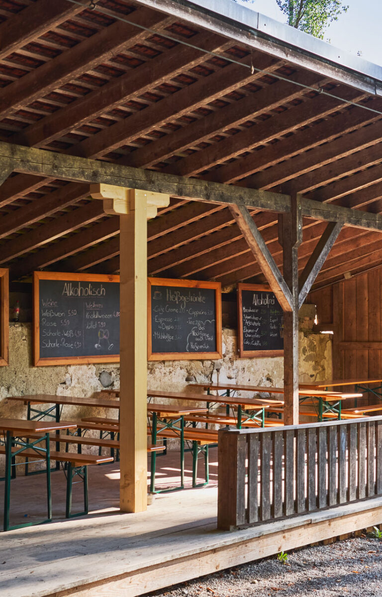 Ein herausstehendes Vordach eines Biergartens. Darunter befinden sich mehrere Biertischgarnituren und das Sonnenlicht strahlt schön herein.