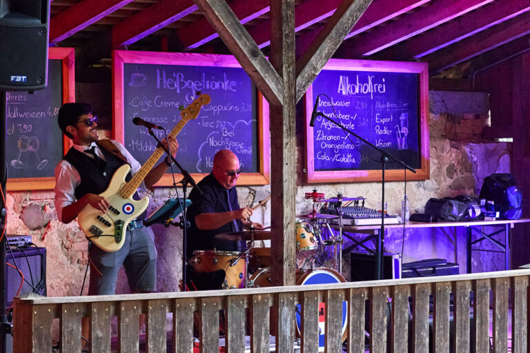 Unter dem Vordach des Biergartens spielt eine Band. Man sieht enen Gitarristen und eine Schlagzeuger, die eine Sonnenbrille targen, lachen und voller Elan spielen. Dahinter erkennt man Effektlichter.