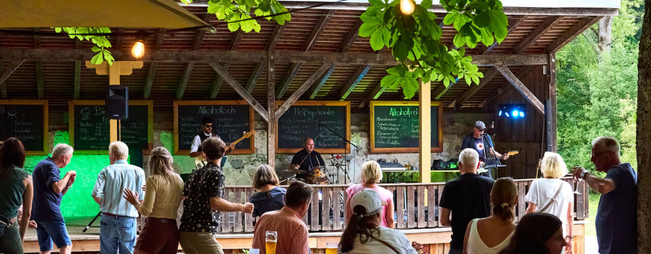 Man sieht wie eine Band unter einem Vordach spielt. E sind drei Musiker, hinter ihnen buntes Licht und vor ihnen viele Menschen die fröhlich tanzen.