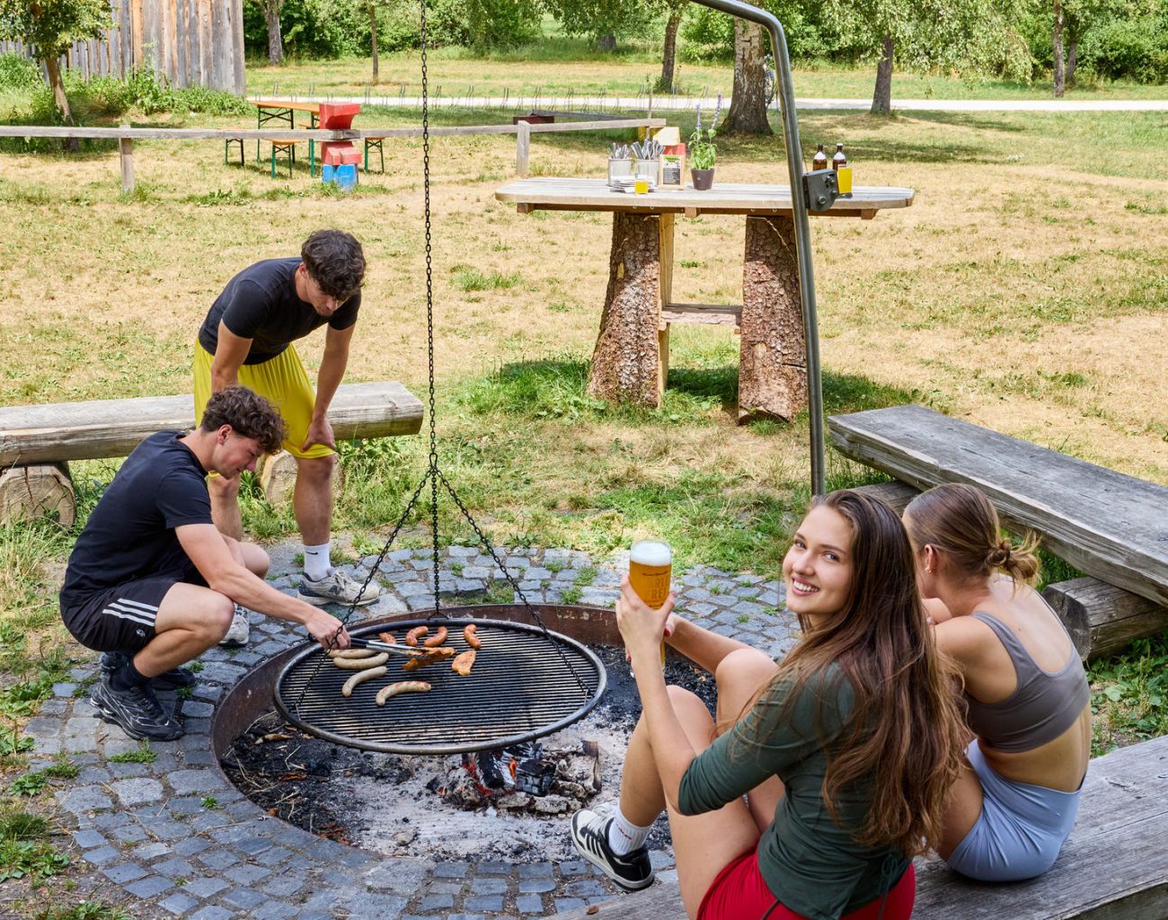 Vier junge Menschen sitzen um eine Grillstelle herum. Die zwei Jungs kümemrn sich um das Grillgut auf dem Rost, die zwei jungen Damen sitzen derweil auf einer Bank. Eines davon lächelt über die Schulter zur Kamera.