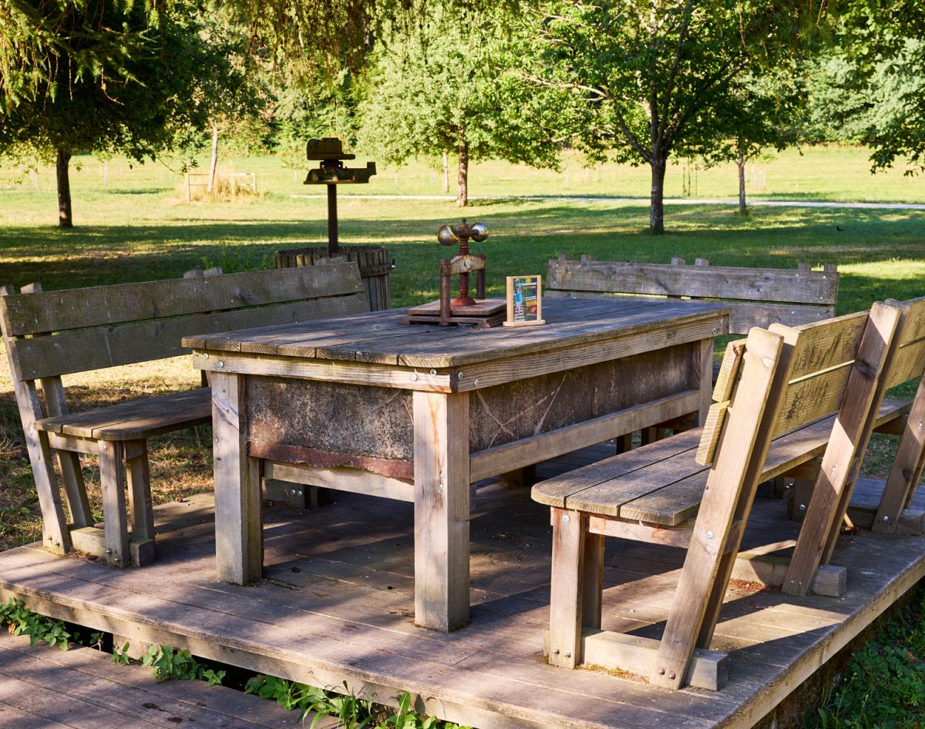 In einer morgendlichen Atmosphäre mit ersten Sonnenstrahlen, steht ein massiver Holztisch im Biergarten mit festinstallierten Holzbänken herum.